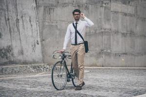 disfrutando del estilo de vida urbano. joven confiado con anteojos mirando hacia otro lado y sosteniendo la mano en su bicicleta mientras camina al aire libre foto