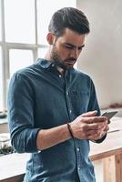 Quick message to client.  Handsome young man holding smart phone and looking at it while standing in the office photo