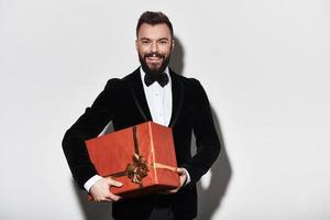 I have something for you. Handsome young man in full suit carrying a gift box and smiling while standing against grey background photo