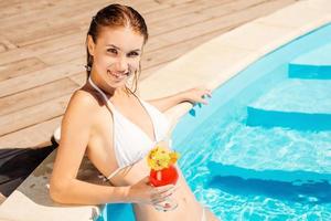 Enjoying her leisure time at the pool. Gorgeous young woman in white bikini holding cocktail and smiling while standing at the pool photo