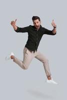 Man in mid-air. Full length of playful young man gesturing and smiling while jumping against grey background photo