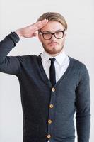 Yes sir Handsome young man in glasses holding hand near head and looking at camera while standing against grey background photo