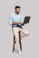 Working day. Full length of good looking young man smiling and using computer while sitting on stool against grey background photo