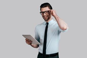 Always ready to help.  Handsome young man in white shirt and tie holding digital while and adjusting his eyeglasses while standing against grey background photo