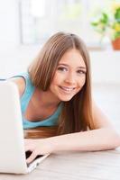 adolescente despreocupado. alegre joven adolescente usando computadora y sonriendo mientras se acuesta en el suelo en su apartamento foto