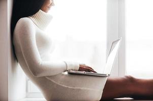 All the world in one touch. Cropped image of attractive young African woman in warm sweater working on laptop while sitting on the window sill photo