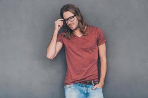 Enjoying his casual style. Handsome young man in casual wear adjusting his eyeglasses and looking thoughtful while standing against grey background photo