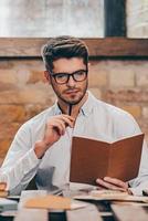 Finding the perfect solution. Handsome young man holding note pad and looking at it while sitting at his working place photo