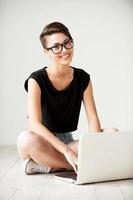 belleza con laptop. hermosa joven mujer de pelo corto trabajando en una laptop y sonriendo a la cámara mientras se sienta en el suelo foto