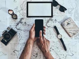 Ready to see the world. Close up top view of man using smart phone with sunglasses, photo camera, compass, watch and passport lying on map around