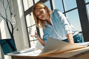 Beautiful young woman looking at blueprint and smiling while working in the office photo