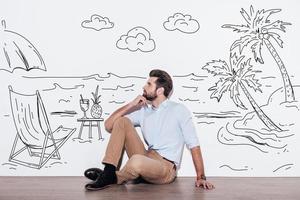 Dreaming about vacation. Young handsome man keeping hand on chin and looking away while sitting on the floor with illustration of resort in the background photo