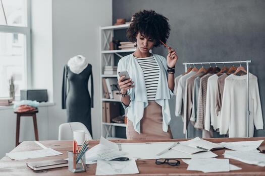 escribiendo un mensaje de negocios. una joven africana pensativa mirando un teléfono inteligente y tocándose la cabeza con un lápiz mientras estaba de pie en su taller con ropa colgada en el fondo foto