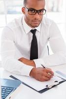 Confident businessman at work. Top view of confident young African man in shirt and tie writing something at clipboard and looking at camera while sitting at his working place photo