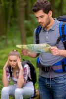 atrapado en algún lugar del bosque. un joven pensativo con mochila examinando un mapa en el bosque mientras una mujer se sienta en el fondo y sostiene la cabeza en la mano foto