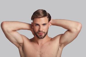 Just perfect. Thoughtful young man looking at camera and keeping hands behind head while standing against grey background photo