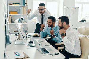 Finally success. Group of young modern men in formalwear smiling and gesturing while working in the office photo