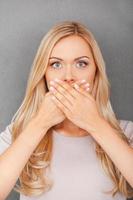 Less information. Surprised young blond hair woman covering her mouth and staring at camera while standing against grey background photo