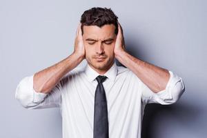 I need silence. Frustrated young man in shirt and tie covering ears with hands and keeping eyes closed while standing against grey background photo