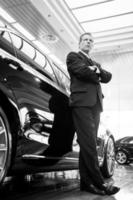 Only luxury cars. Black and white image of full length confident grey hair man in formalwear leaning at the car and looking away photo