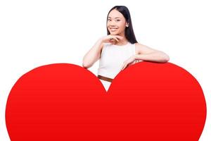 I love you as much Beautiful young Asian woman in pretty dress looking at camera leaning on huge heart shaped valentine card and smiling while standing against white background photo
