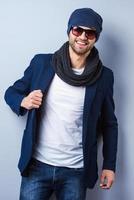 Confident in his perfect style. Handsome young stylish man in sunglasses and hat adjusting his jacket and looking at camera while standing against grey background photo
