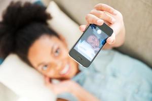 selfie perezoso. vista superior de una atractiva joven africana con auriculares escuchando música y sonriendo mientras está tumbada en el sofá de casa foto