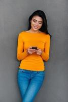 Quick message to friend. Attractive young woman holding smart phone looking at it with smile while standing against grey background photo