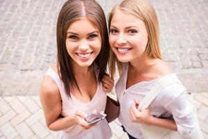 Cheerful friends. Top view of two beautiful young women smiling at camera while standing close to each other outdoors photo