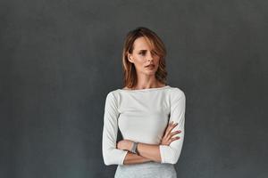 Young expert. Thoughtful young woman in smart casual wear keeping arms crossed and looking away while standing against grey background photo