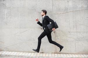 apresurándose a las nuevas metas. joven feliz con ropa formal sosteniendo un maletín mientras corre frente a la pared de hormigón foto