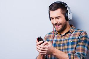 Love this music. Handsome young man in headphones holding mobile phone and smiling while standing against grey background photo