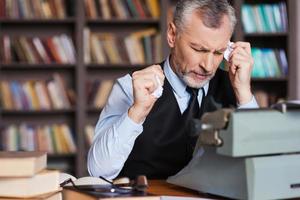 That is all wrong Frustrated grey hair senior man in formalwear sitting at the typewriter and holding pieces of paper in his hands with bookshelf in the background photo