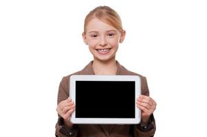 Copy space at her tablet. Cheerful little girl in formalwear showing her digital tablet and smiling while standing isolated on white photo