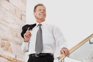 Confident businessman. Cheerful mature man in formalwear smiling while moving down by staircase photo