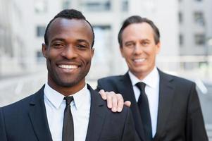 Business support. Confident senior man in formalwear holding hand on shoulder of young African man that looking at camera and smiling photo