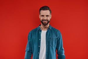 Handsome young man in casual clothing smiling and looking at camera while standing against red background photo