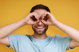 Portrait curious young man in casual clothing gesturing eyeglasses and smiling while standing against yellow background photo