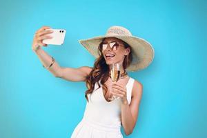 Beautiful young smiling woman in elegant hat holding champagne flute and making selfie photo