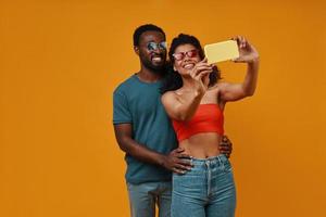 hermosa joven pareja africana sonriendo y tomando selfie mientras está de pie contra el fondo amarillo foto