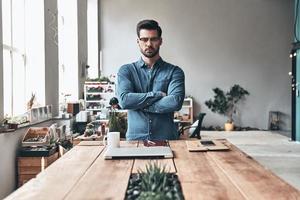 Pure confidence. Handsome young man looking a camera and keeping arms crossed while standing in the creative working space photo