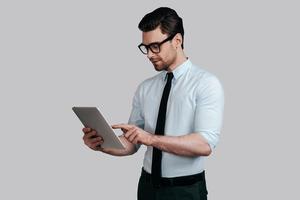 utilizando tecnologías modernas. un joven apuesto con camisa blanca y corbata trabajando en su tableta digital mientras se enfrenta a un fondo gris foto