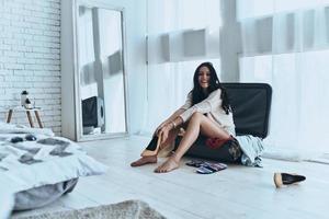 Getting ready to vacation.  Beautiful young woman smiling and looking at camera while sitting on the open suitcase at home photo