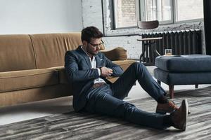 Looking just perfect. Handsome young man in full suit adjusting his sleeve while sitting on the floor at home photo