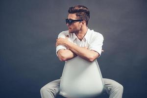 Doing it in style. Confident young handsome man in sunglasses looking away while sitting in chair against grey background photo