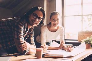 Young and successful. Cheerful young man and woman looking at camera and smiling while leaning at office desk with blueprint laying on it photo