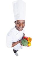 Healthy food is in his hands. Top view of cheerful young African chef in white uniform holding colorful vegetables while standing against white background photo