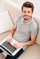 Working at home. Top view of young man working on laptop and looking at camera while sitting on sofa photo