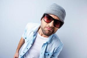 Am I cool enough Top view of handsome young man in hat and sunglasses looking at camera and grimacing while standing against grey background photo