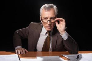 Trying to remember you. Serious senior man in formalwear sitting at his working place and adjusting his glasses while isolated on black background photo